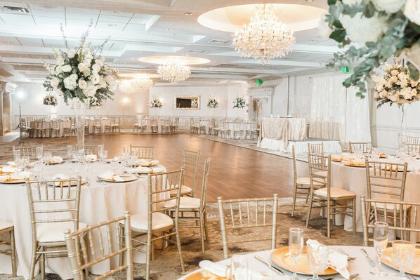 A large, elegantly decorated banquet hall with round tables, gold chairs, and hanging floral arrangements.