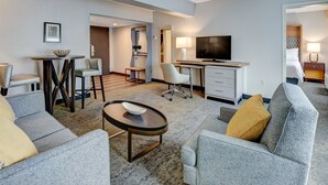 A modern hotel room with a grey sofa, a small round table, a desk with a TV, and a bed with a headboard.