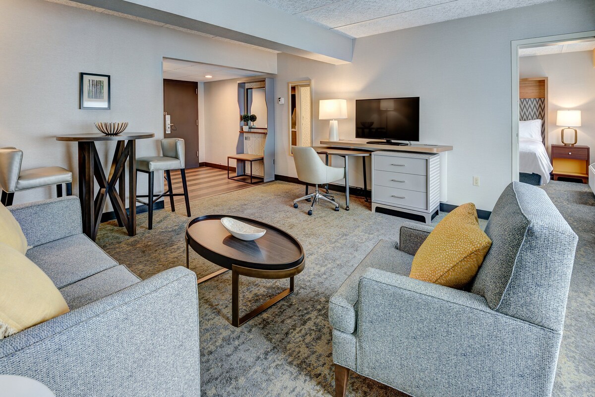 A modern hotel room with a grey sofa, a small round table, a desk with a TV, and a bed with a headboard.