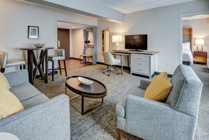 Una habitación de hotel moderna con un sofá gris, una mesita redonda pequeña, un escritorio con televisor y una cama con cabecera.