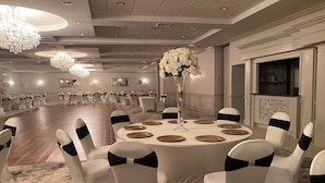 A banquet hall with round tables, white chairs with black sashes, a tall vase with white flowers, chandeliers, and a fireplace.