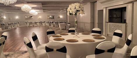 Uma sala de banquetes com mesas redondas, cadeiras brancas com fitas pretas, um vaso alto com flores brancas, lustres e uma lareira.