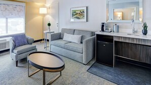 A modern hotel room with a grey sofa, a small round table, a desk with a sink, and a framed artwork on the wall.