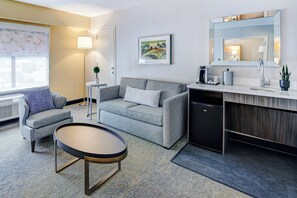 A modern hotel room with a grey sofa, a small round table, a desk with a sink, and a framed artwork on the wall.