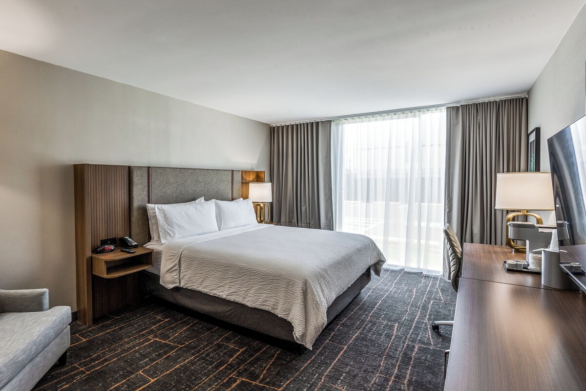 A hotel room with a large bed, a desk with a lamp, a chair, and a bedside table with a phone and remote control.