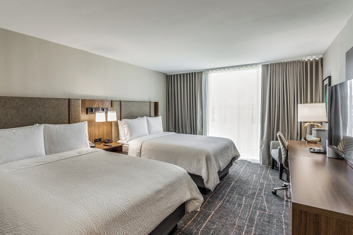 A hotel room with two beds, a desk with a TV, and a window with curtains.