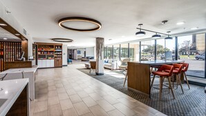 A modern hotel lobby with a reception desk, a snack bar, seating area, and large windows offering a view of the outdoors.
