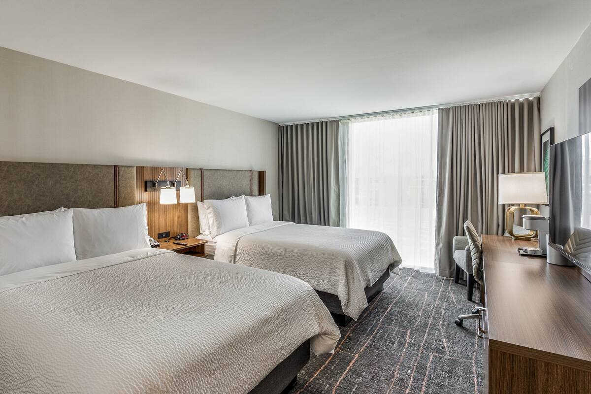 Une chambre d’hôtel avec deux lits, un bureau avec une télévision et une fenêtre avec des rideaux.
