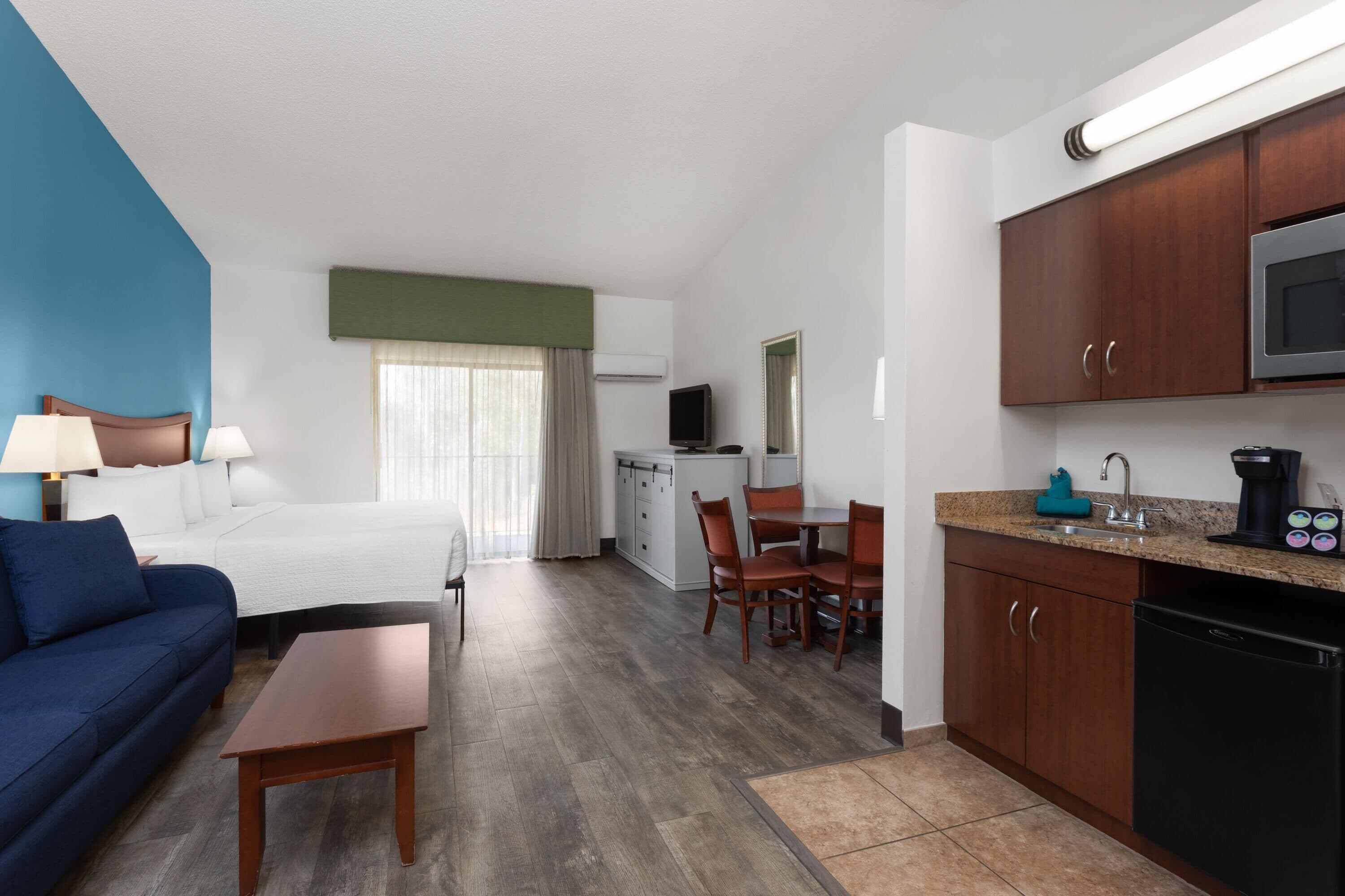 A hotel room with a bed, a blue sofa, a small table, a kitchenette with a microwave, a coffee maker, and a TV.