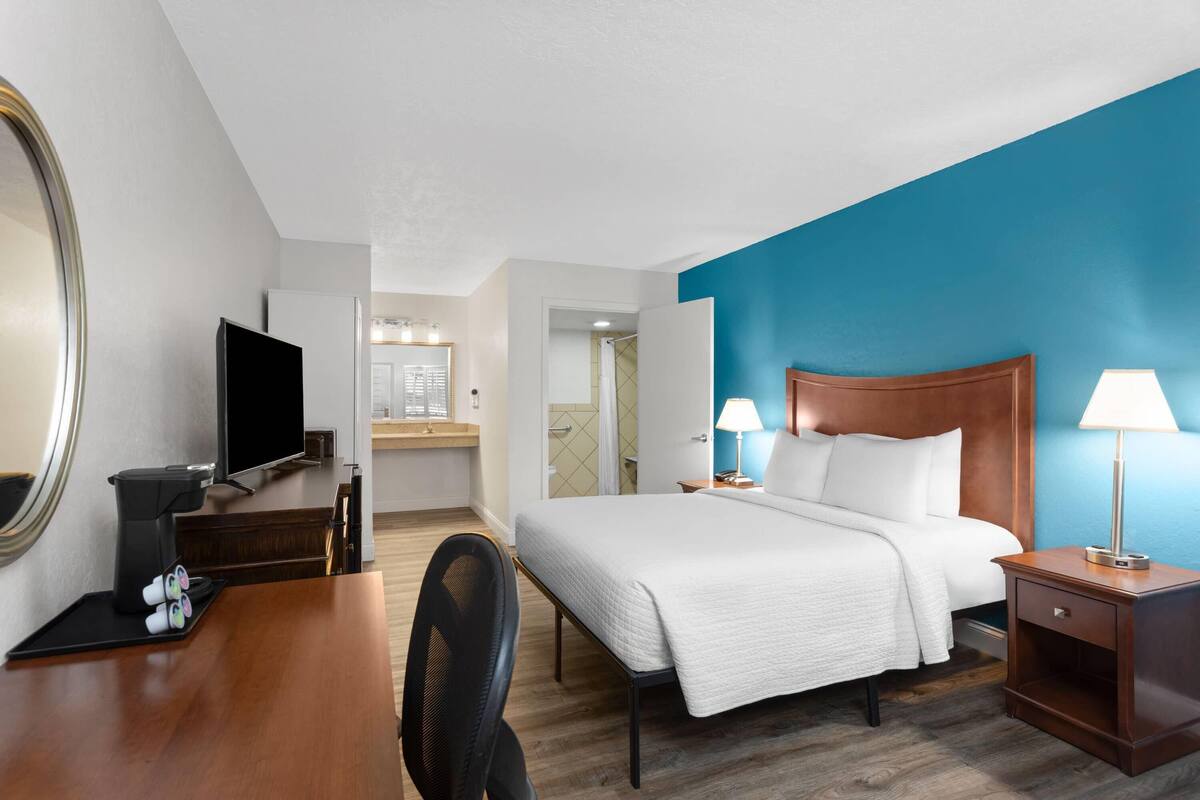 A hotel room with a bed, a desk with a coffee maker, a TV, and a bathroom visible in the background.