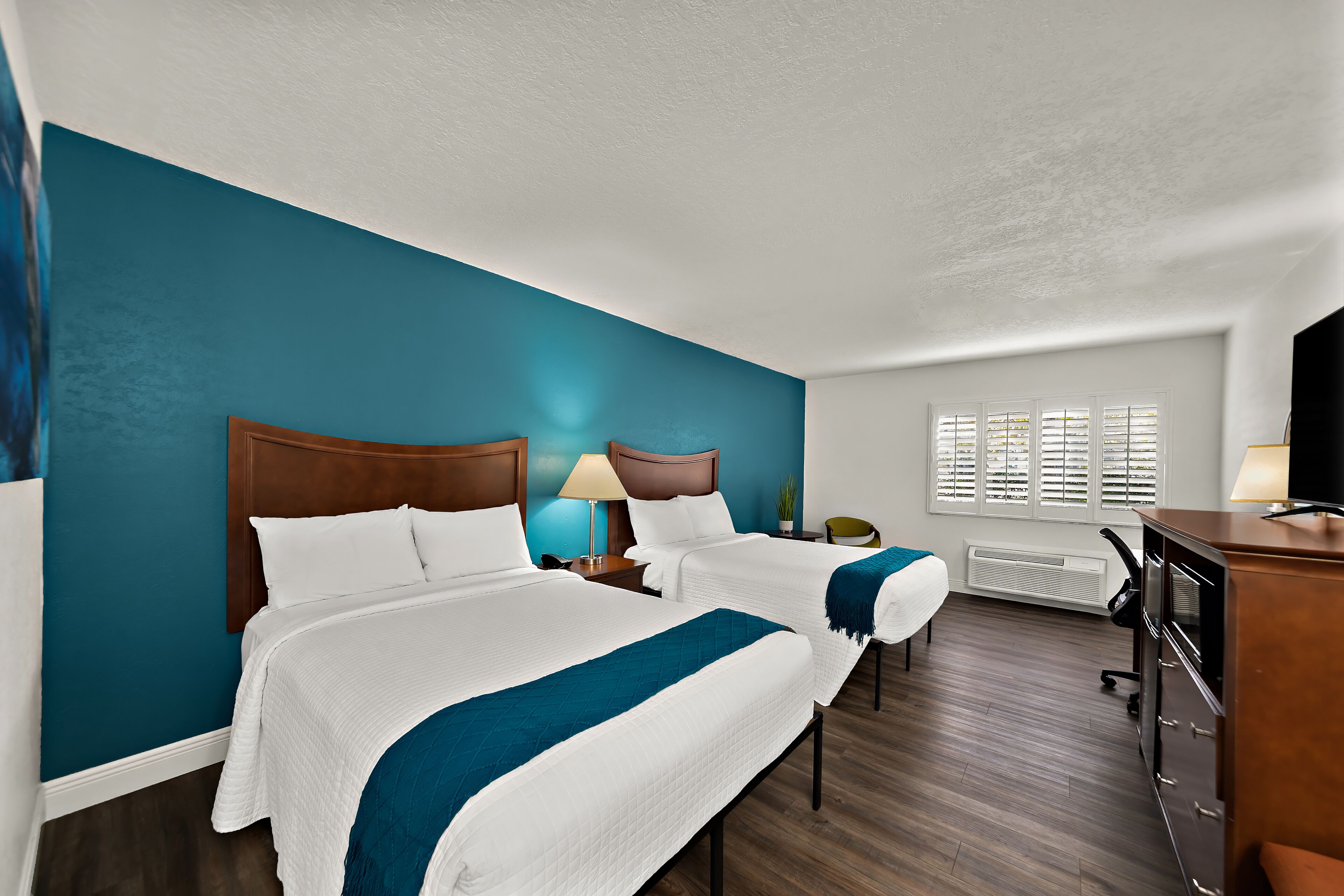 Quarto de hotel com duas camas, uma escrivaninha, uma cadeira, uma televisão e janela com persianas.