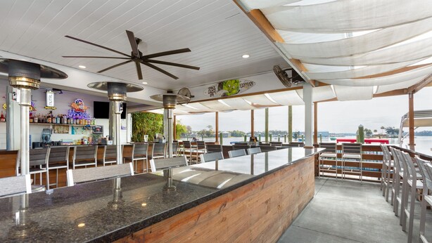 Una zona de bar con mostrador de madera, asientos, ventiladores de techo y vistas al agua.