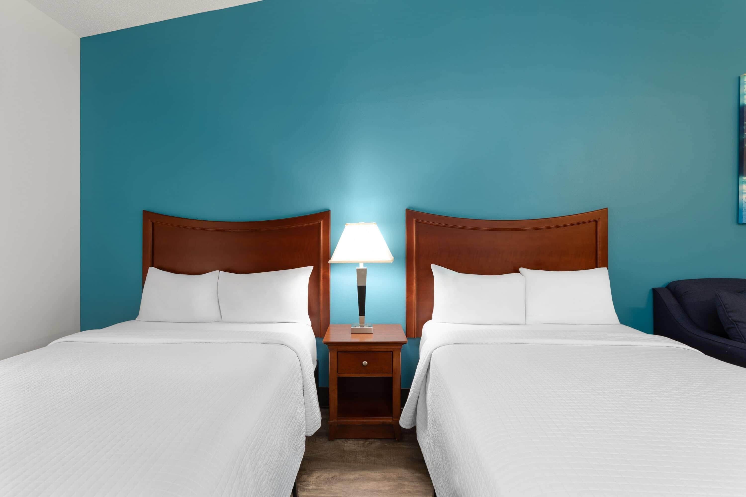 A hotel room with two beds, a nightstand with a lamp, and a dark-colored armchair.