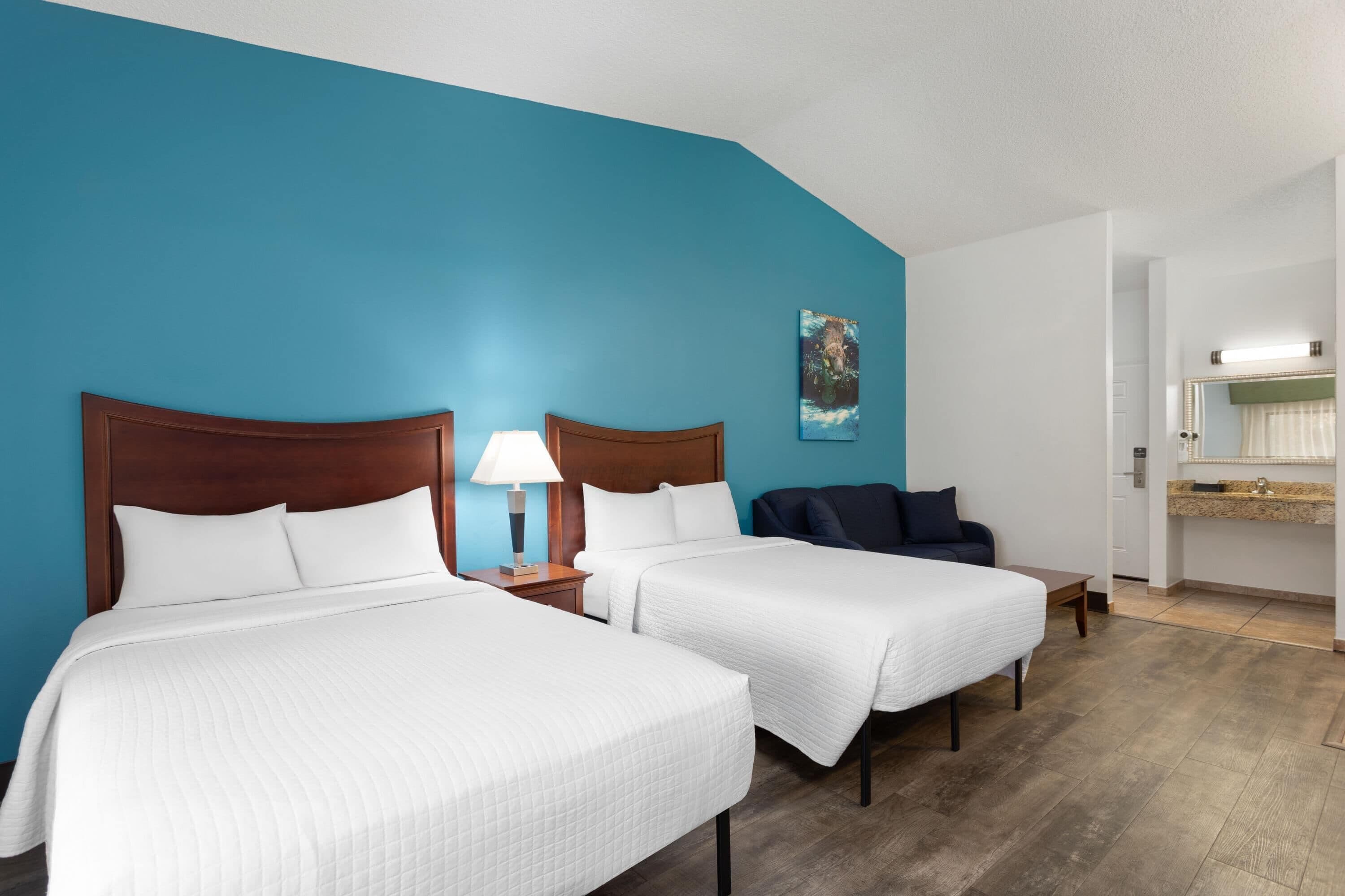 A hotel room with two beds, a nightstand with a lamp, a blue accent wall, a painting, and a bathroom with a vanity and mirror.