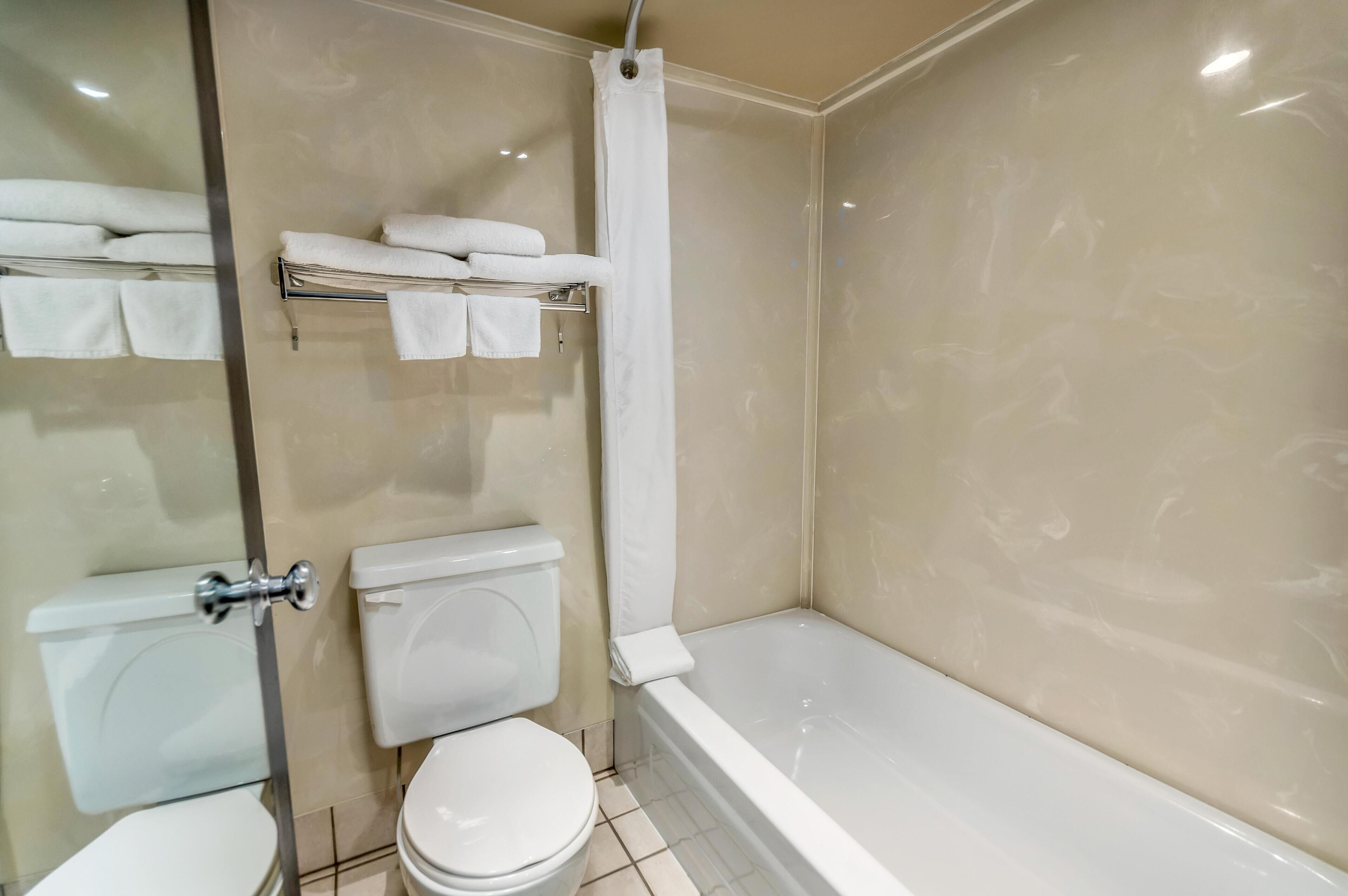 A bathroom with a glass shower enclosure, a toilet, a bathtub, and a shelf with folded towels.