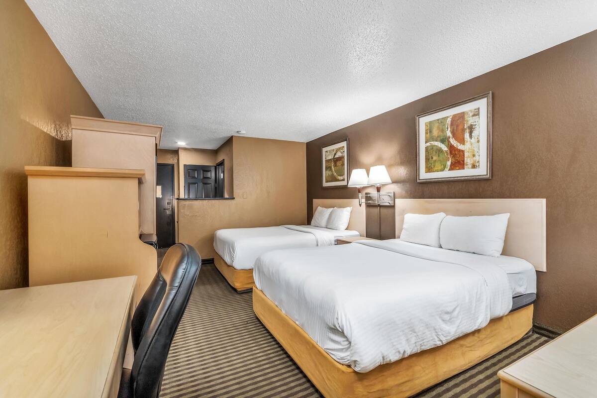 Quarto de hotel com duas camas, uma escrivaninha, uma cadeira, uma luminária e obras de arte nas paredes.