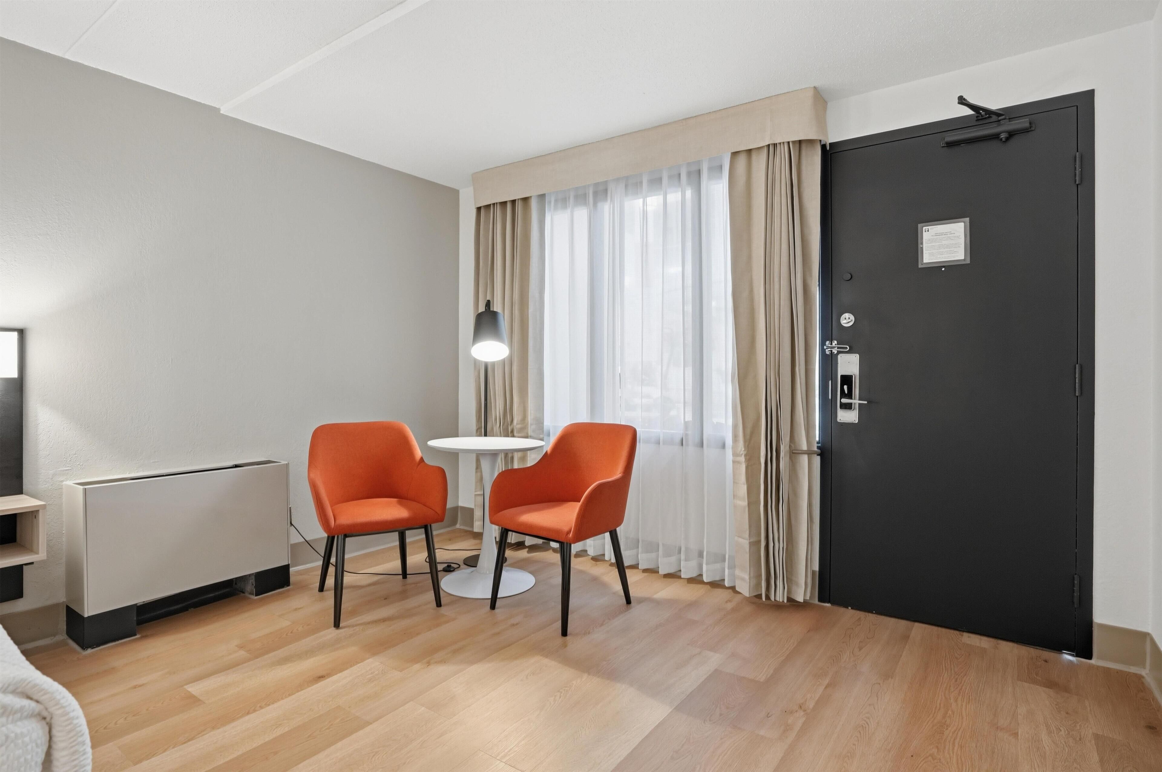 A hotel room with a bed, a radiator, two orange chairs, a small table, a lamp, a black door, and a window with curtains.