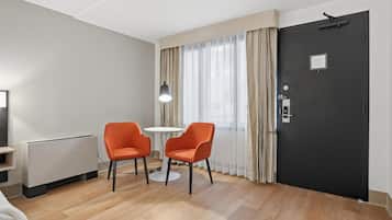 A hotel room with a bed, a radiator, two orange chairs, a small table, a lamp, a black door, and a window with curtains.