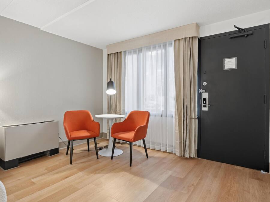 A hotel room with a bed, a radiator, two orange chairs, a small table, a lamp, a black door, and a window with curtains.