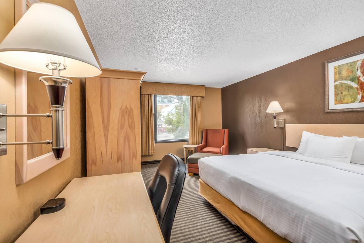 Quarto de hotel com cama, escrivaninha, cadeira, luminária e janela com cortinas.