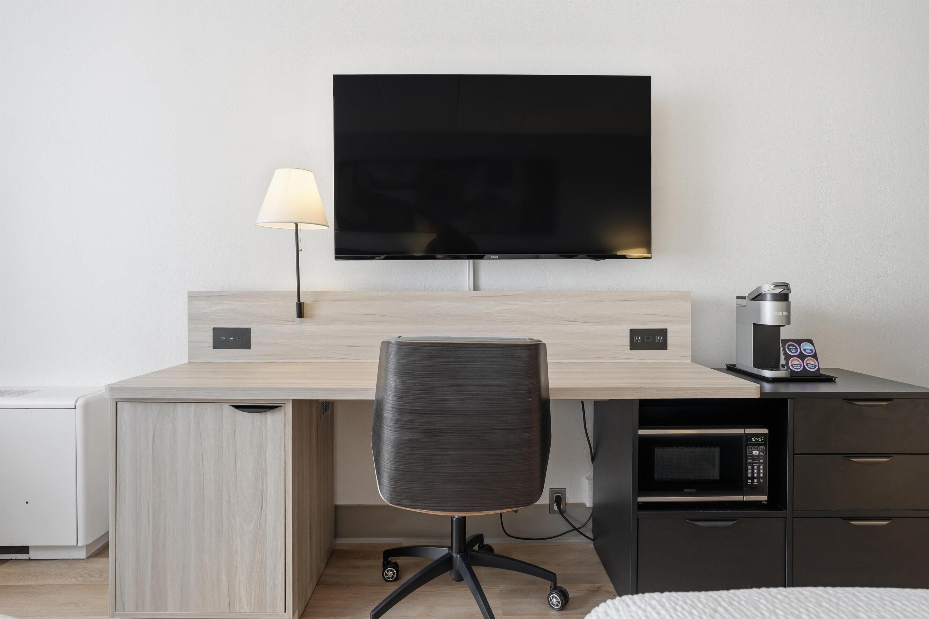 A modern hotel room with a flat-screen TV mounted on the wall, a desk with a lamp, a chair, a coffee machine, and a microwave.
