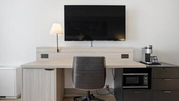 A modern hotel room with a flat-screen TV mounted on the wall, a desk with a lamp, a chair, a coffee machine, and a microwave.