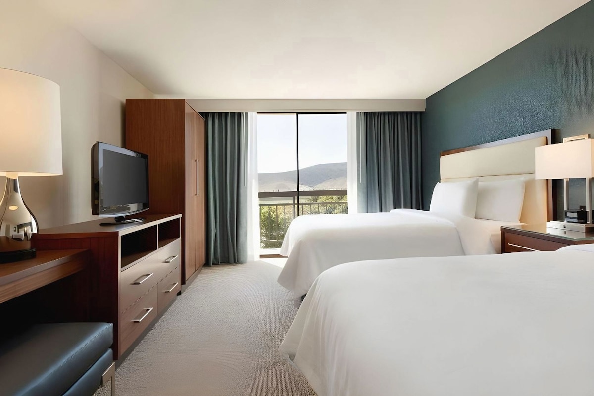 A hotel room with two beds, a TV, a desk with a lamp, and a view of the outdoors.