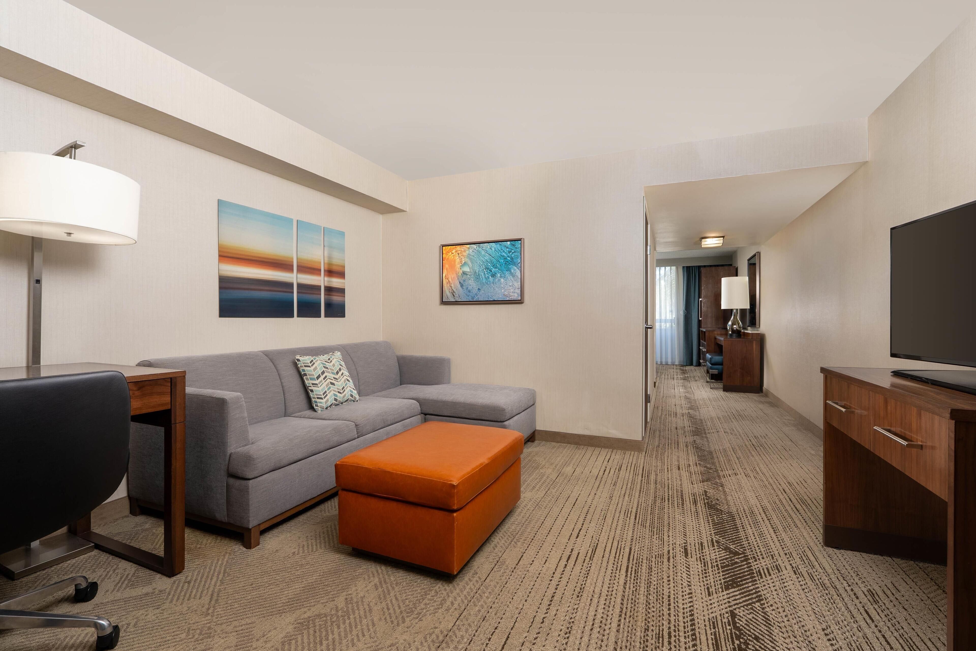 A hotel room with a gray sofa, an orange ottoman, a wooden desk, a TV, and artwork on the walls.