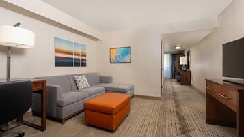 A hotel room with a gray sofa, an orange ottoman, a wooden desk, a TV, and artwork on the walls.