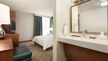 Ein Hotelzimmer mit einem Bett, einem Schreibtisch mit Lampe, einem großen Spiegel und einem Badezimmer mit Waschbecken und Handtuchhalter.