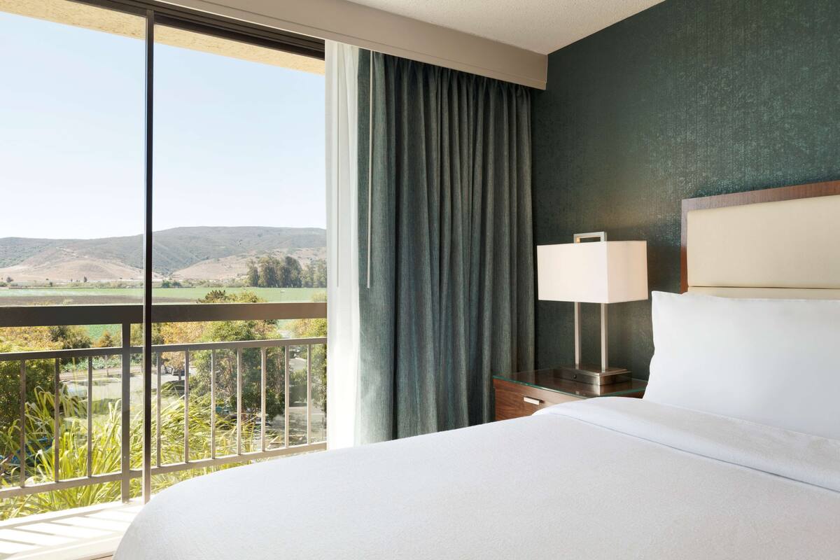 Una habitació d'hotel amb un llit gran, una tauleta de nit amb una làmpada i vistes a un paisatge des del balcó.