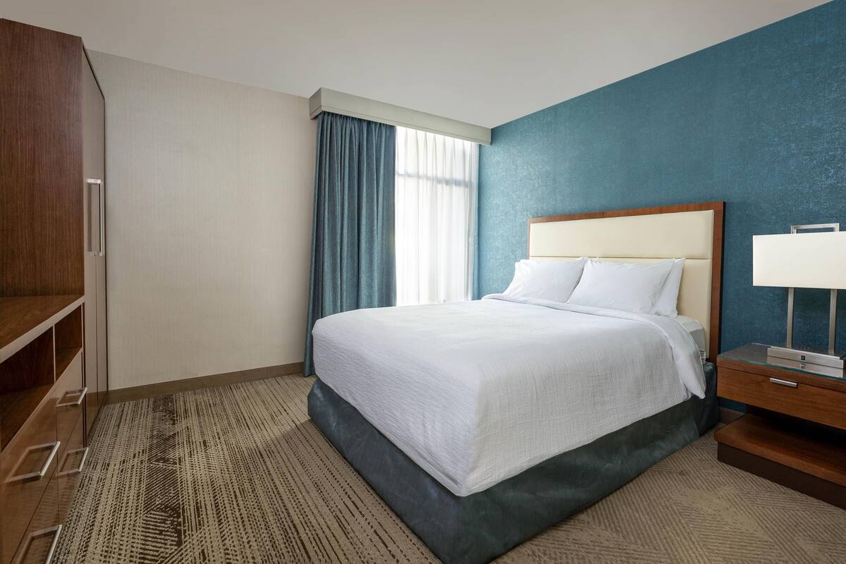 Habitació d'hotel amb un llit gran, un armari de fusta, una taula de nit amb una làmpada i una finestra amb cortines.