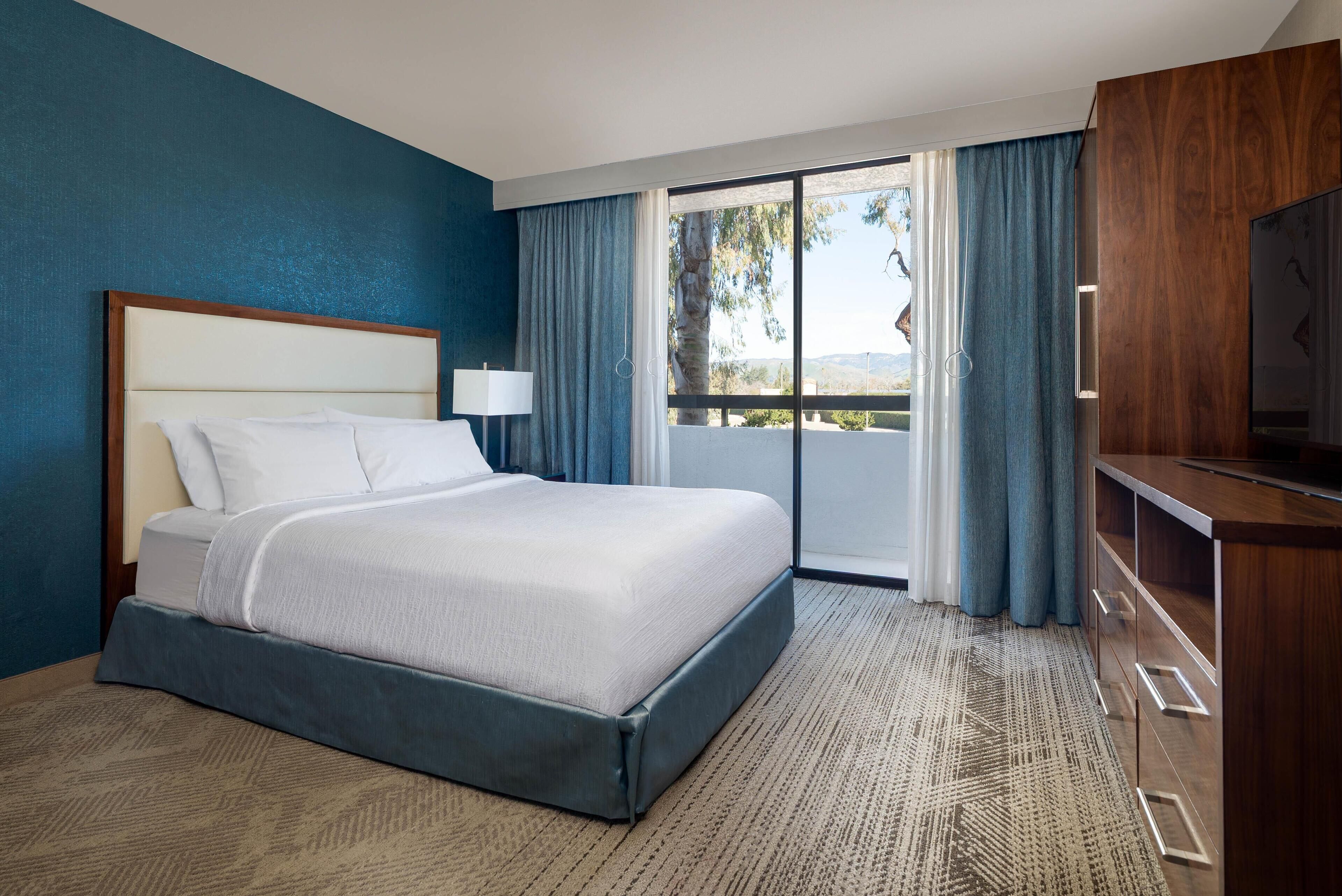 Habitación de hotel con una cama grande, televisor, balcón con cortinas y vistas al exterior.
