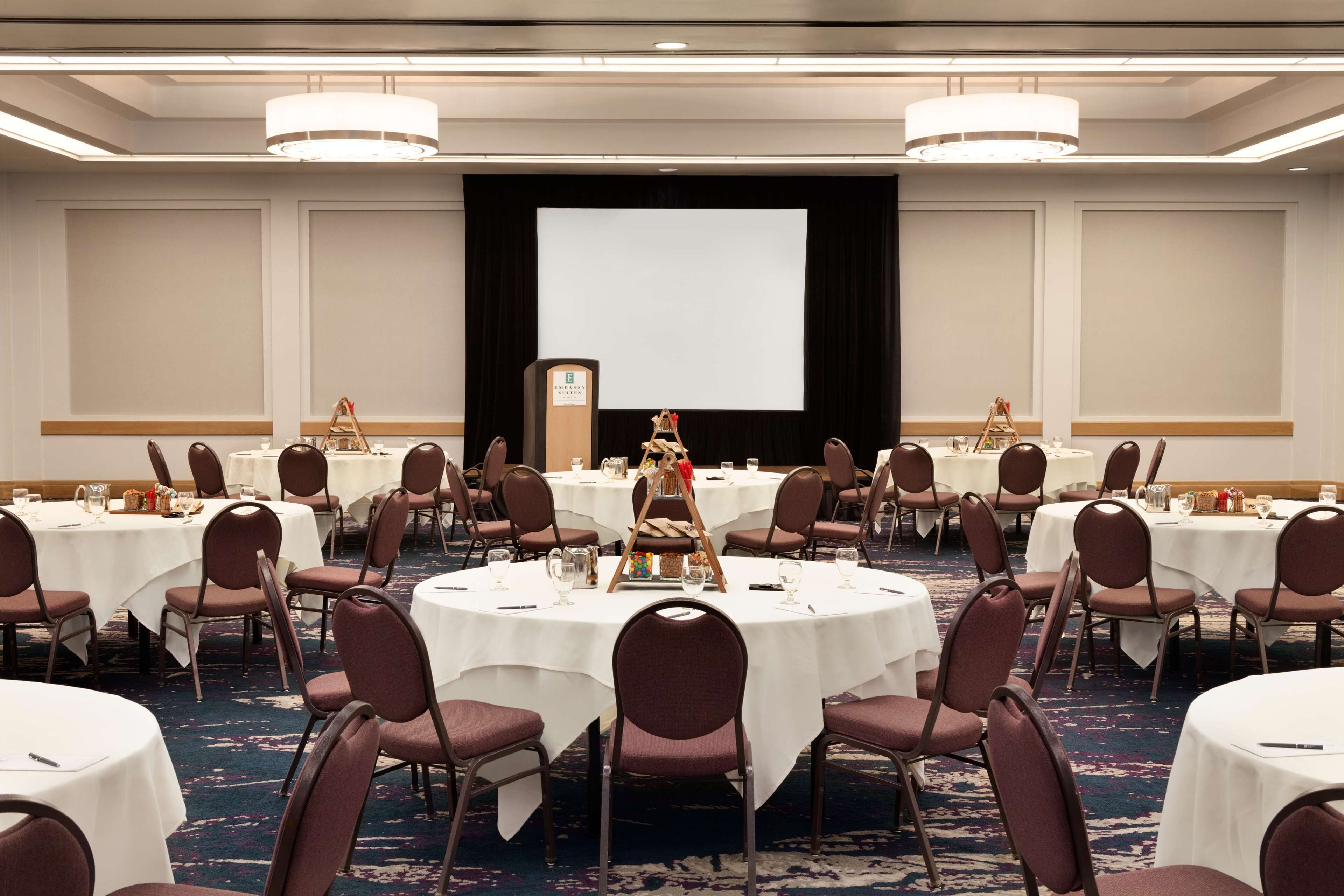 A conference room with round tables, chairs, a projector screen, and a podium.