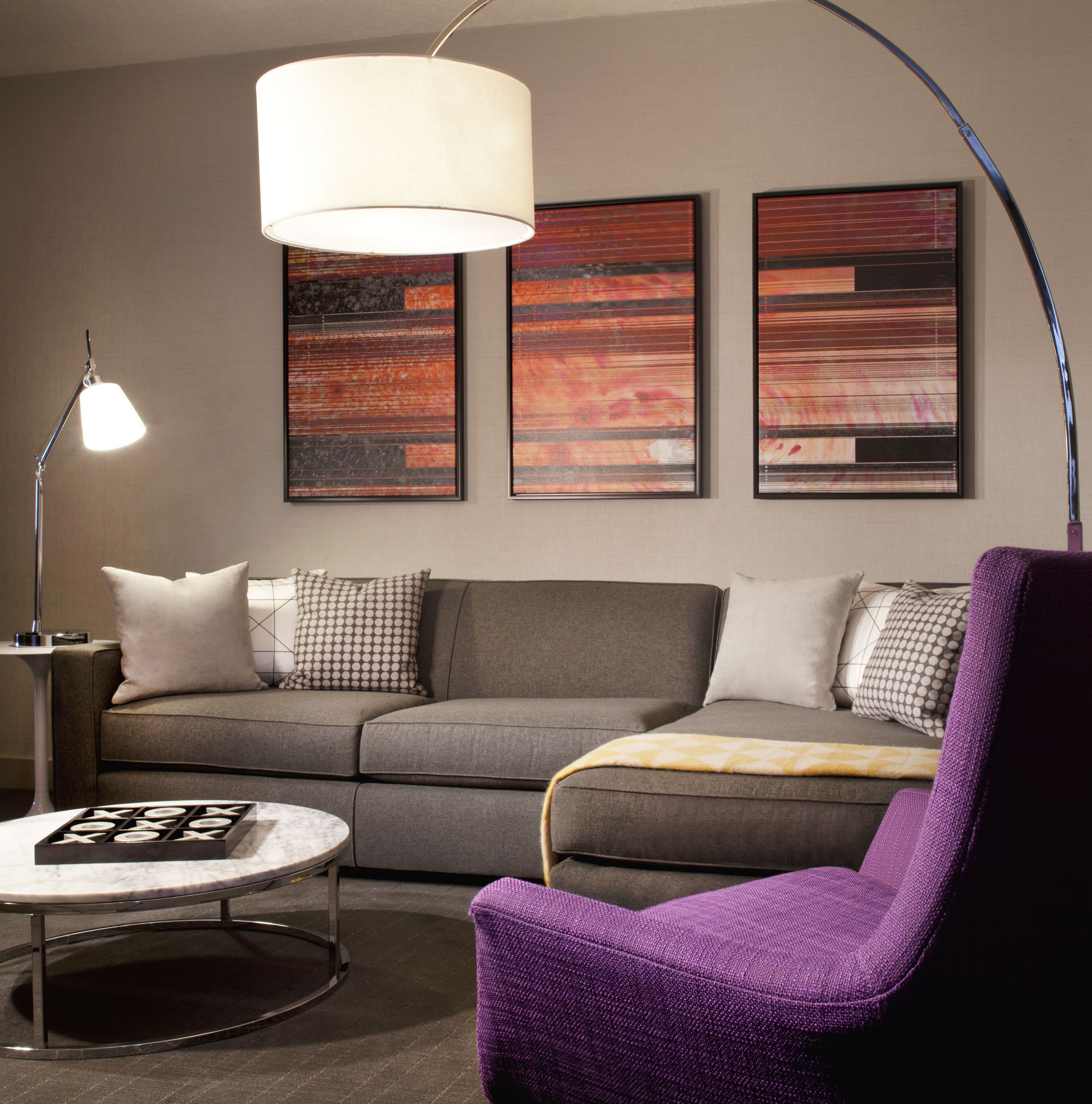 A modern living room with a grey sofa, a purple armchair, a round coffee table, and a floor lamp. There are three abstract paintings on the wall.