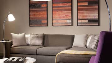 A modern living room with a grey sofa, a purple armchair, a round coffee table, and a floor lamp. There are three abstract paintings on the wall.