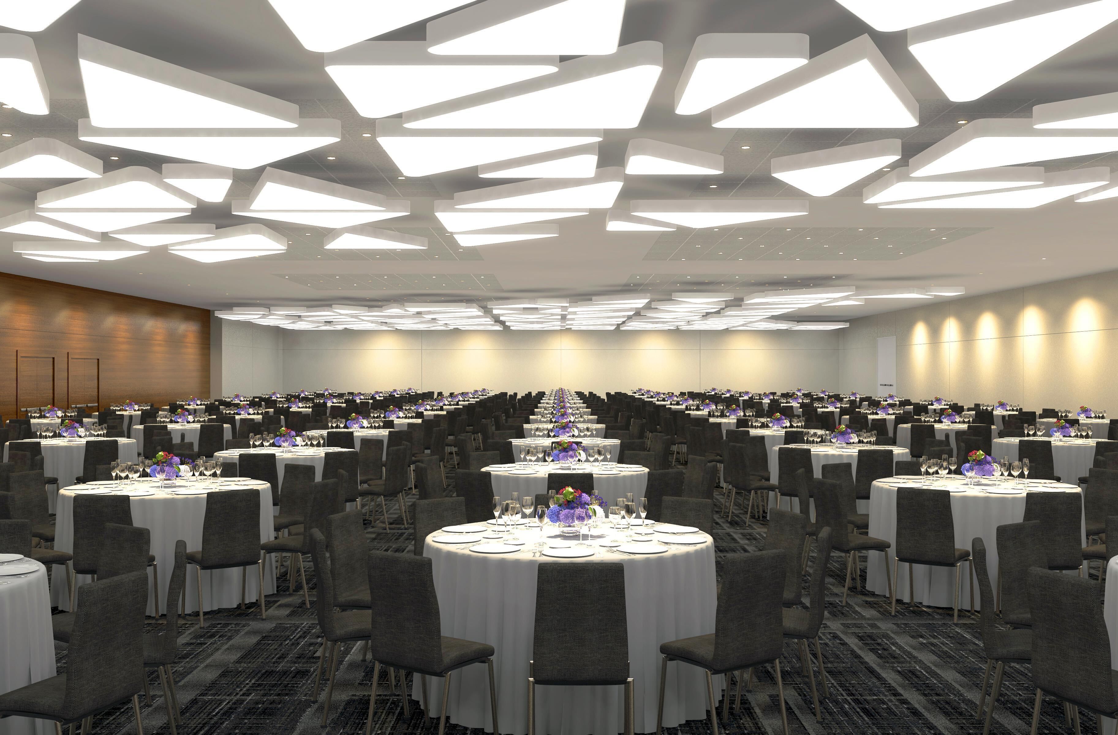 Une vaste salle de banquet dotée de nombreuses tables rondes, chacune recouverte d’une nappe blanche et entourée de chaises, et agrémentée d’un design de plafond moderne.