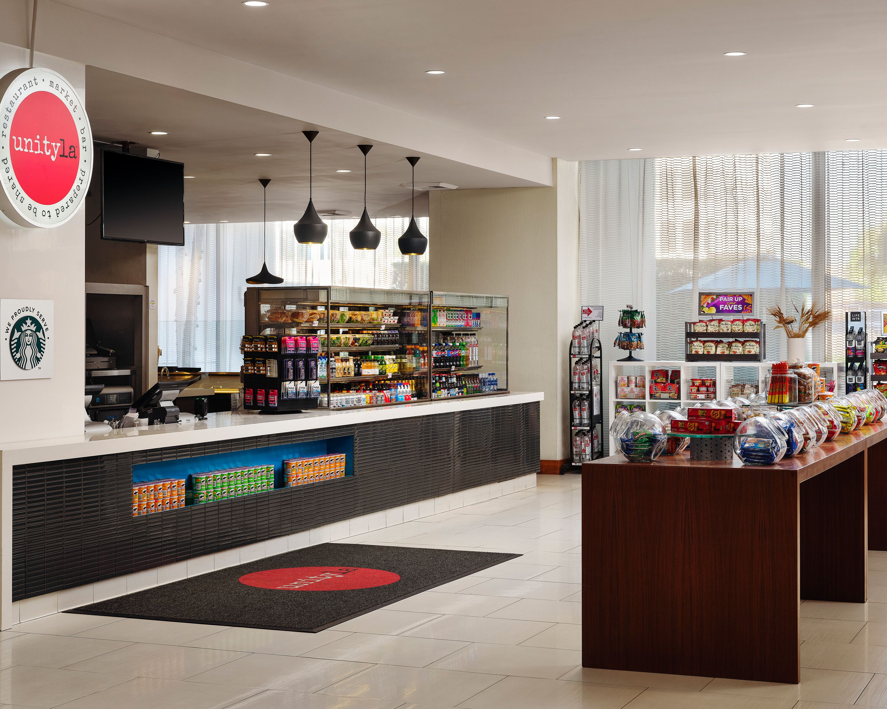 Un espacio comercial moderno con un mostrador, estanterías llenas de snacks y bebidas, y el logotipo de Starbucks en la pared.