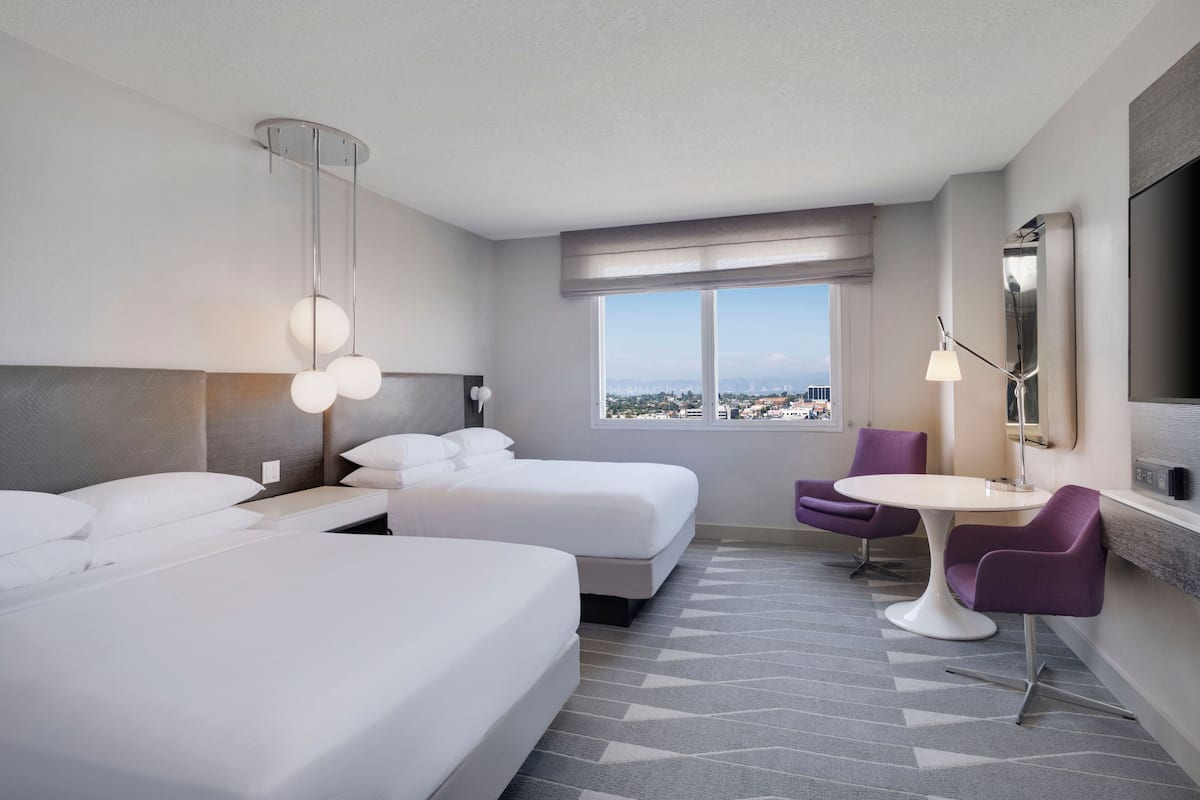 Habitación de hotel con dos camas, una mesa de comedor, una silla, una lámpara y un ventanal con vistas a la ciudad.