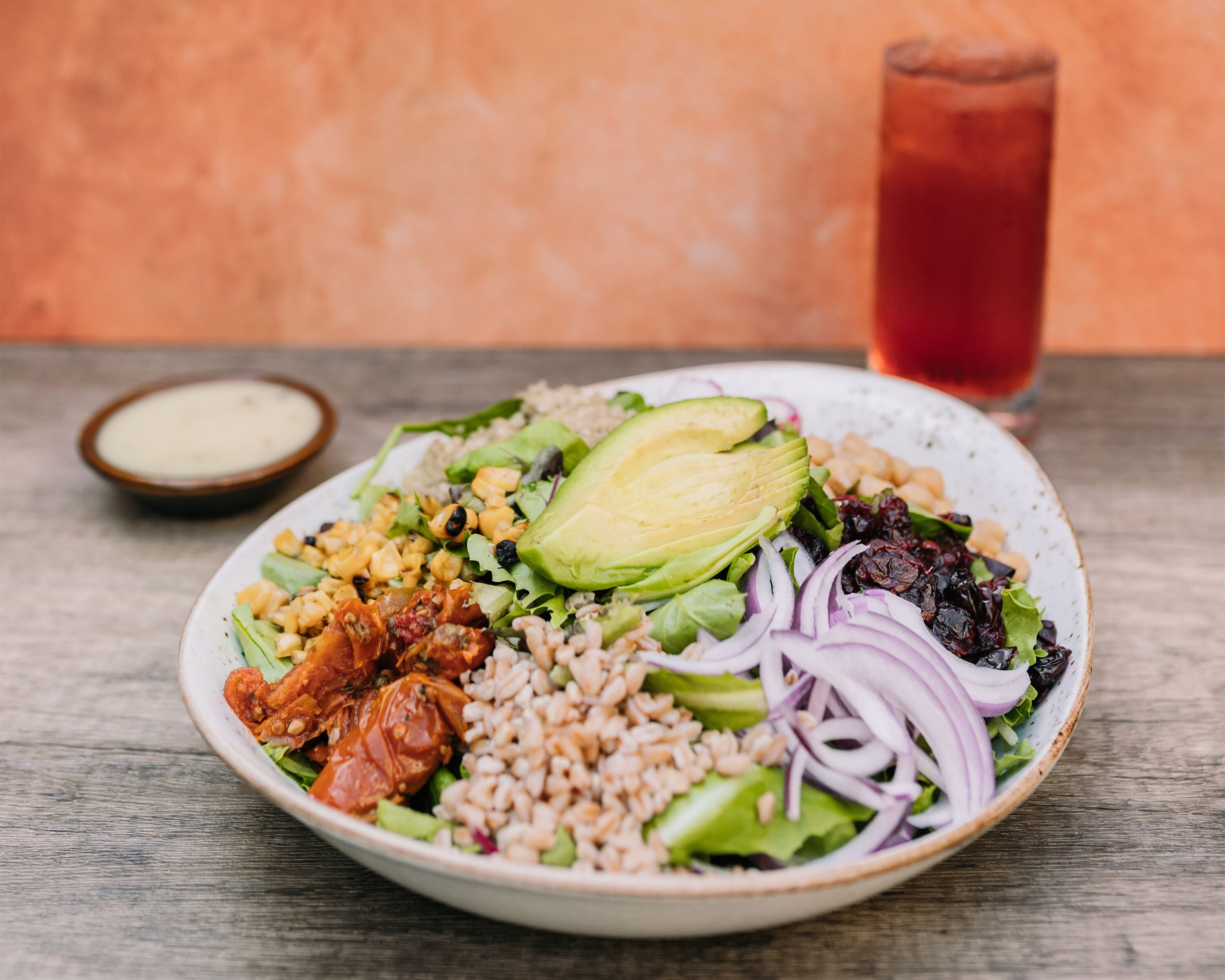 A plate of food with various ingredients including avocado, grains, and vegetables, accompanied by a glass of red beverage and a small bowl of sauce.