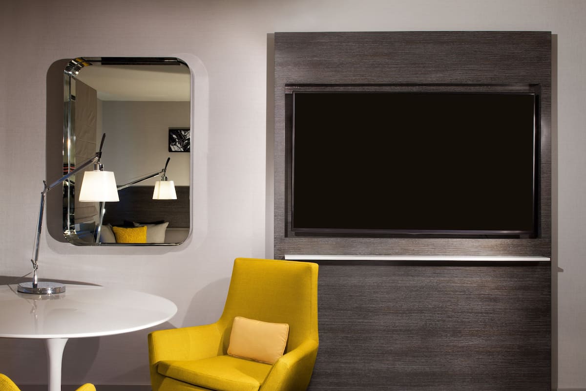 Habitación de hotel con diseño moderno, que incluye un sillón amarillo, una mesa redonda blanca, un espejo grande y un televisor de pantalla plana montado en la pared.