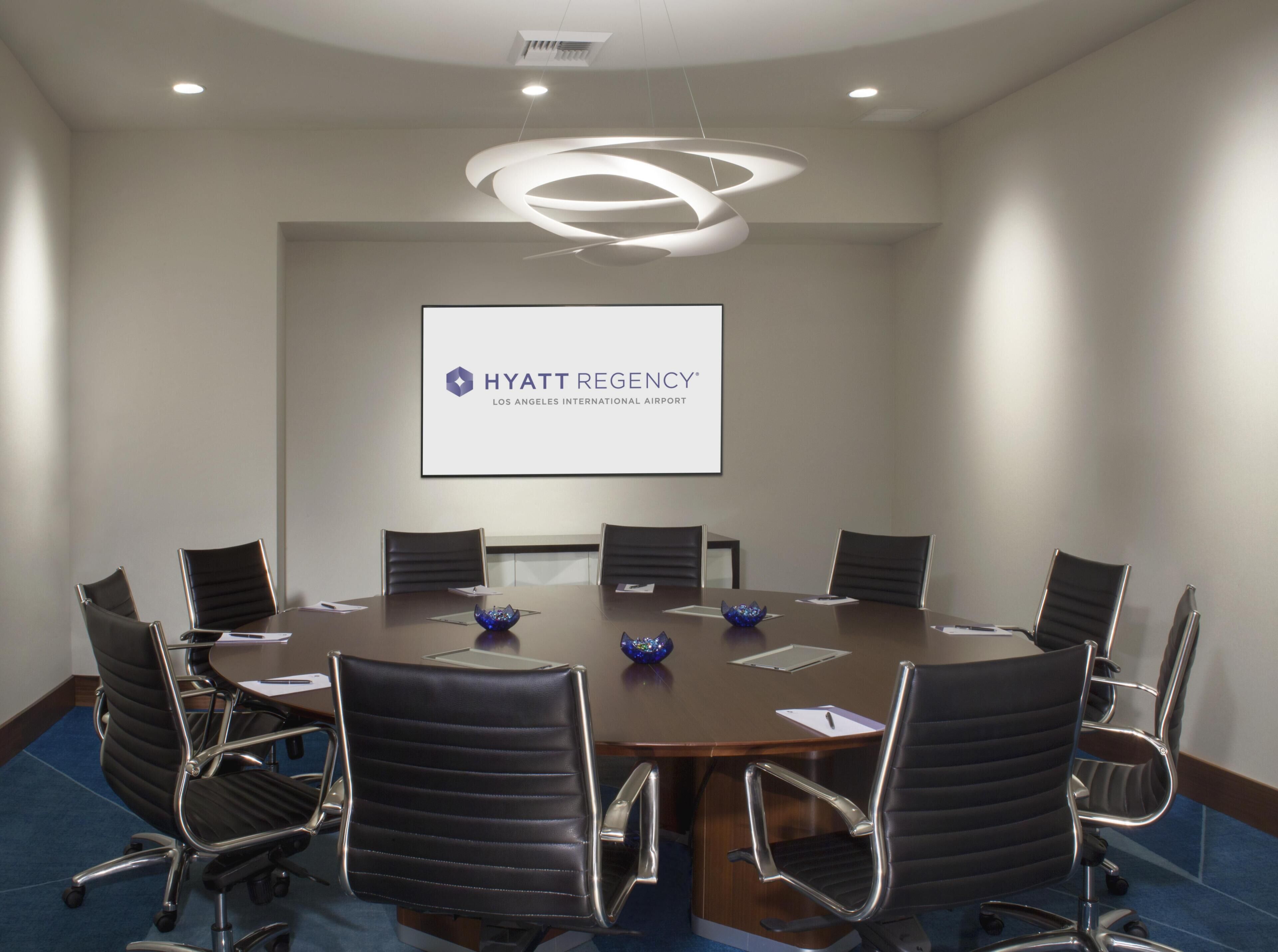 Una sala de conferències amb una taula rodona, cadires negres, una pantalla de projector que mostra 'HYATT REGENCY LOS ANGELES INTERNATIONAL AIRPORT' i una làmpada de sostre moderna.