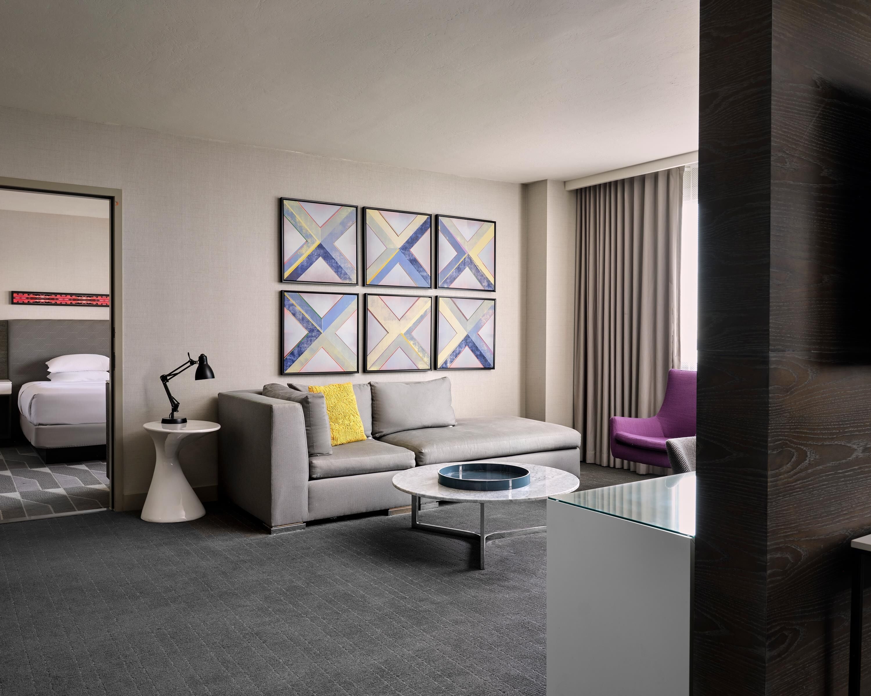 Une chambre d’hôtel moderne avec un canapé gris, une table basse ronde et un fauteuil violet. Des tableaux abstraits ornent le mur, et un lit avec du linge de maison blanc se trouve au fond de la pièce.