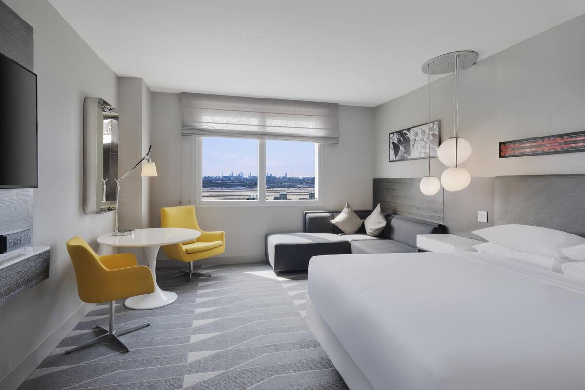 Una habitación de hotel moderna con cama, sofá, mesa de comedor con sillas, televisión y un gran ventanal con vistas a la ciudad.