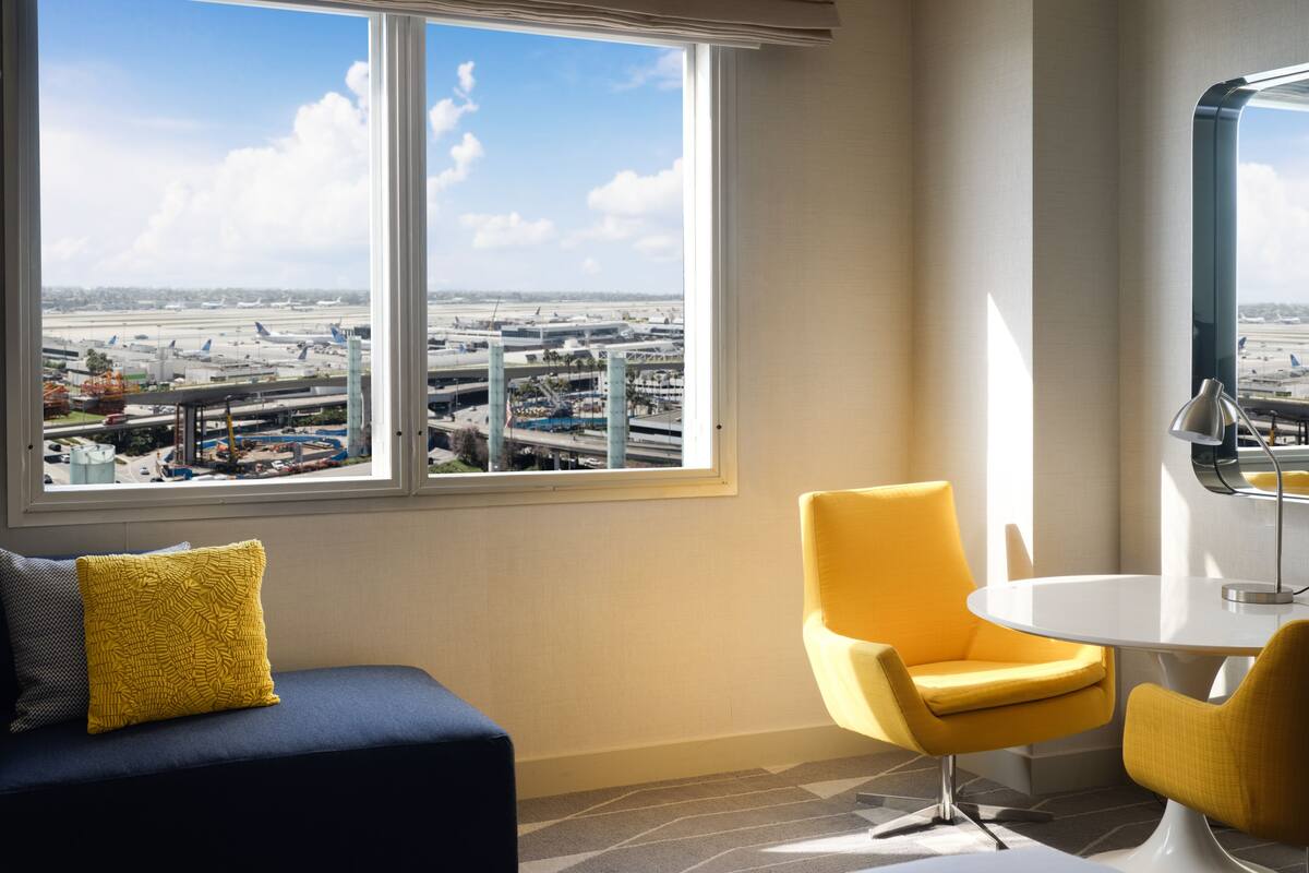 Una habitación con un amplio ventanal con vistas a un aeropuerto, un sofá azul con un cojín amarillo, un sillón amarillo y una mesa blanca.