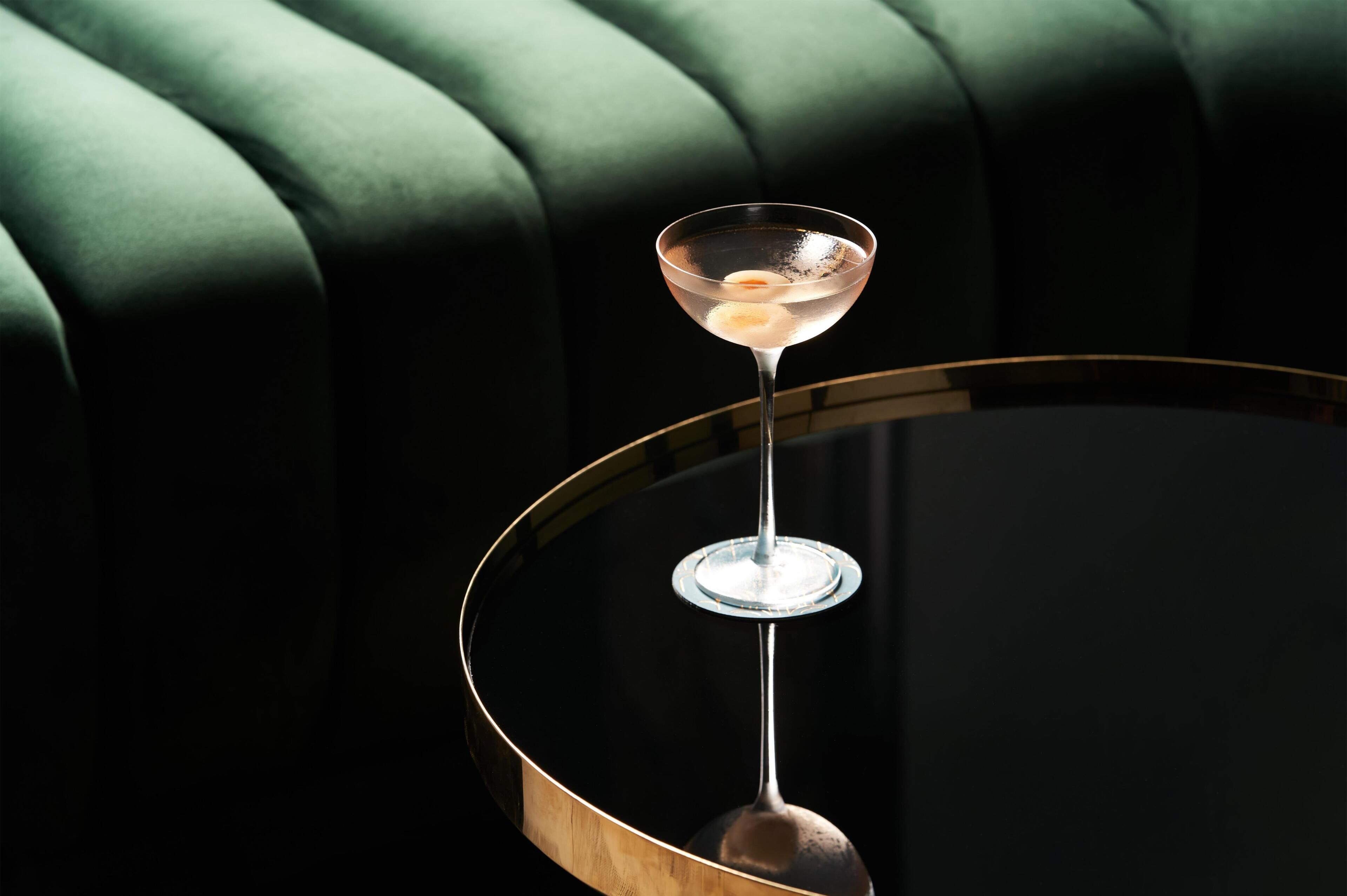 A martini glass on a round table with a gold rim, set against a dark green upholstered chair.