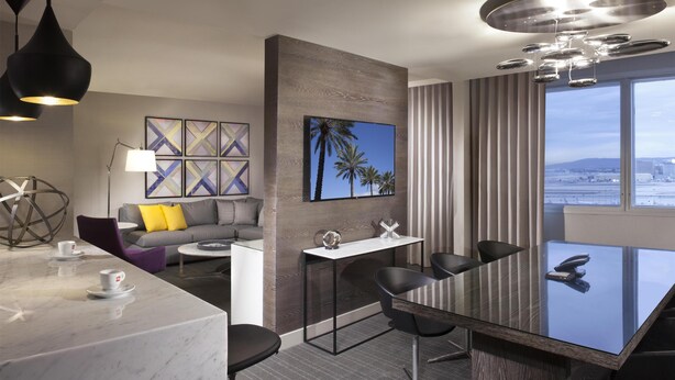 Una habitación de hotel moderna con comedor, sofá con cojines, un ventanal con vistas a la ciudad y una lámpara de techo.