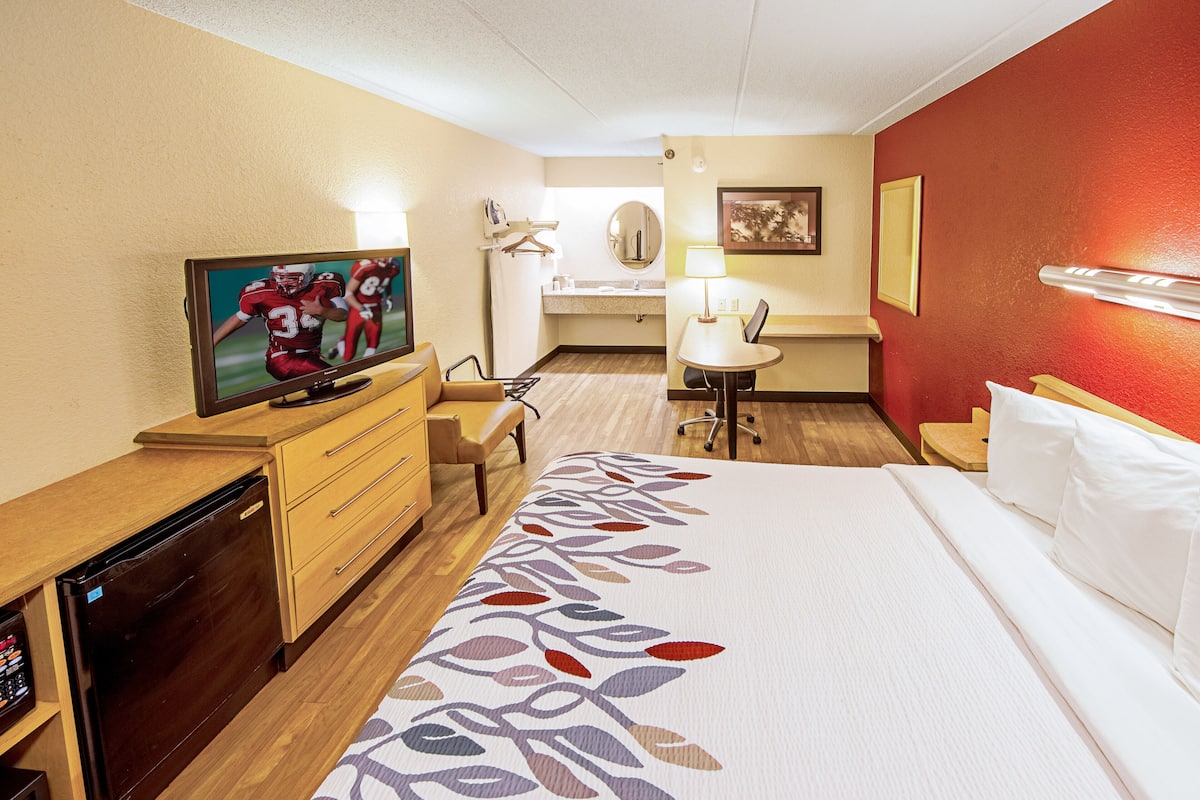 A hotel room with a bed, a TV, a desk, a chair, a small table, and a bathroom visible through an open door.