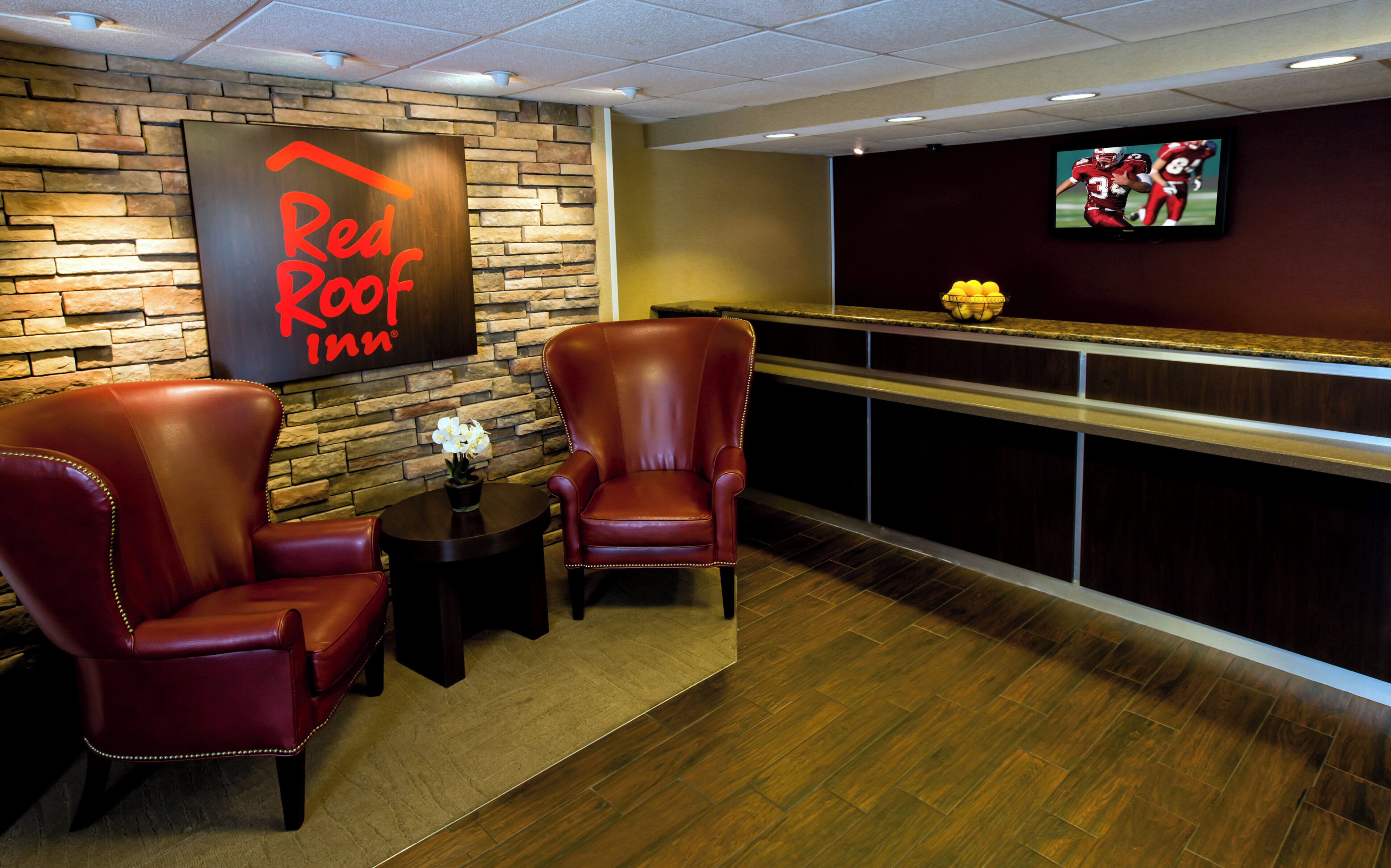 A Red Roof Inn lobby with leather chairs, a small table with a vase, a fruit bowl on the counter, and a TV mounted on the wall.