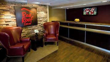 A Red Roof Inn lobby with leather chairs, a small table with a vase, a fruit bowl on the counter, and a TV mounted on the wall.